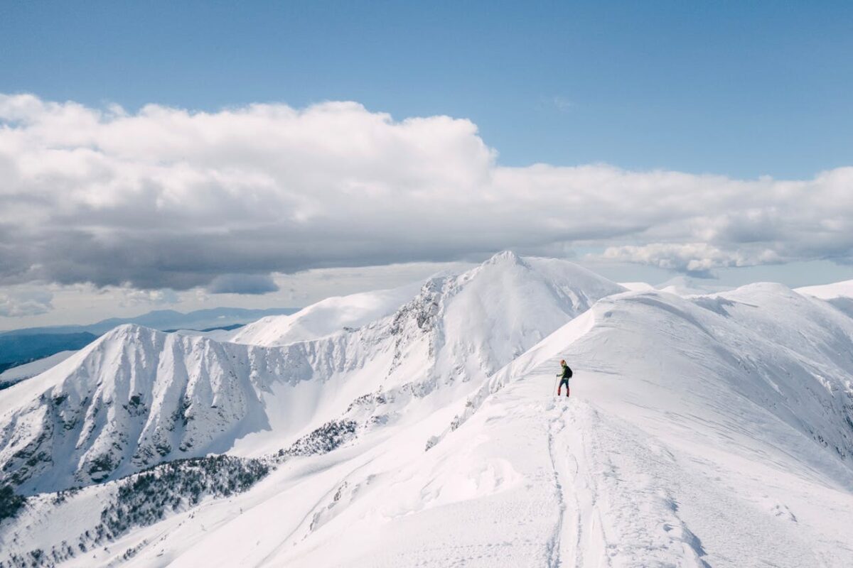 A lone hiker explores a snowy mountain ridge under a bright sky, with breathtaking views.