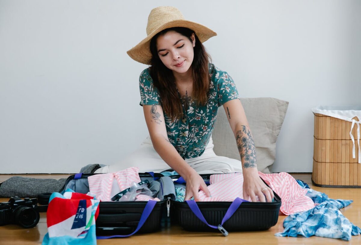 Woman packing suitcase with clothes for a vacation, wearing straw hat and smiling.